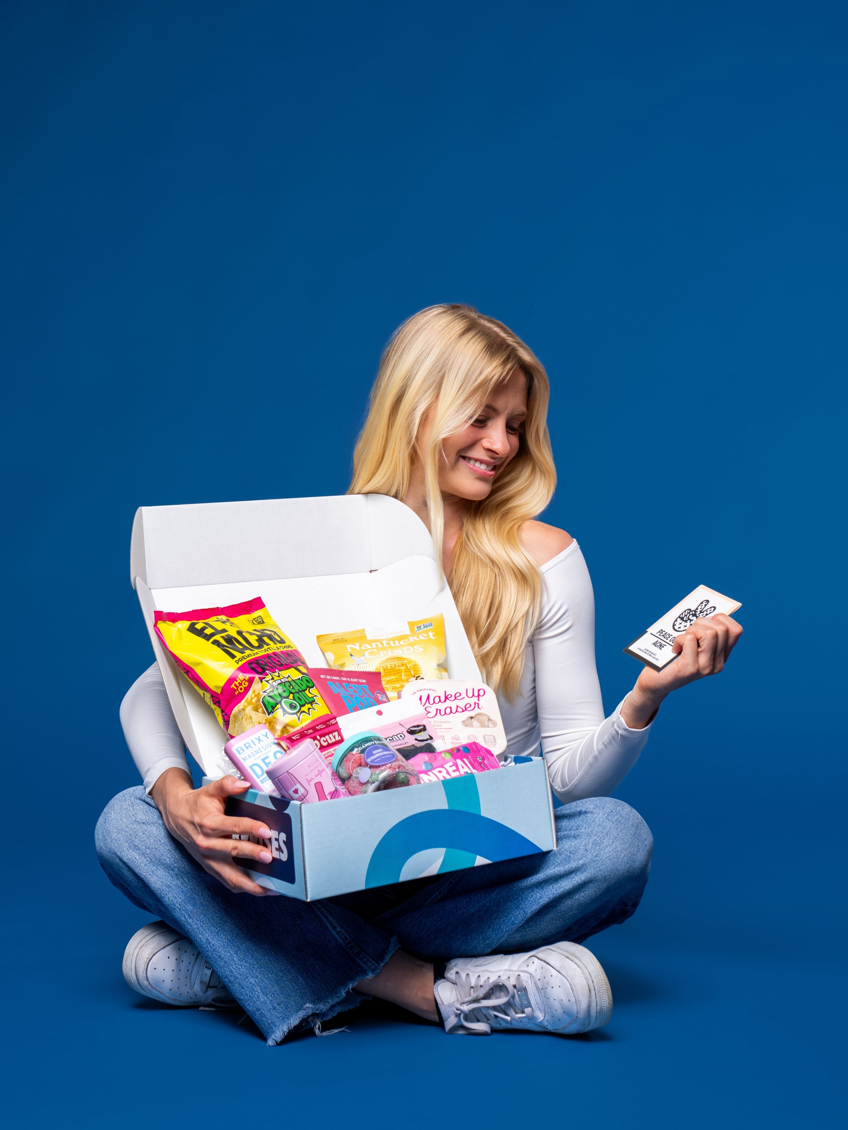 A blonde college student sits cross-legged holding an open DormDrop care package box