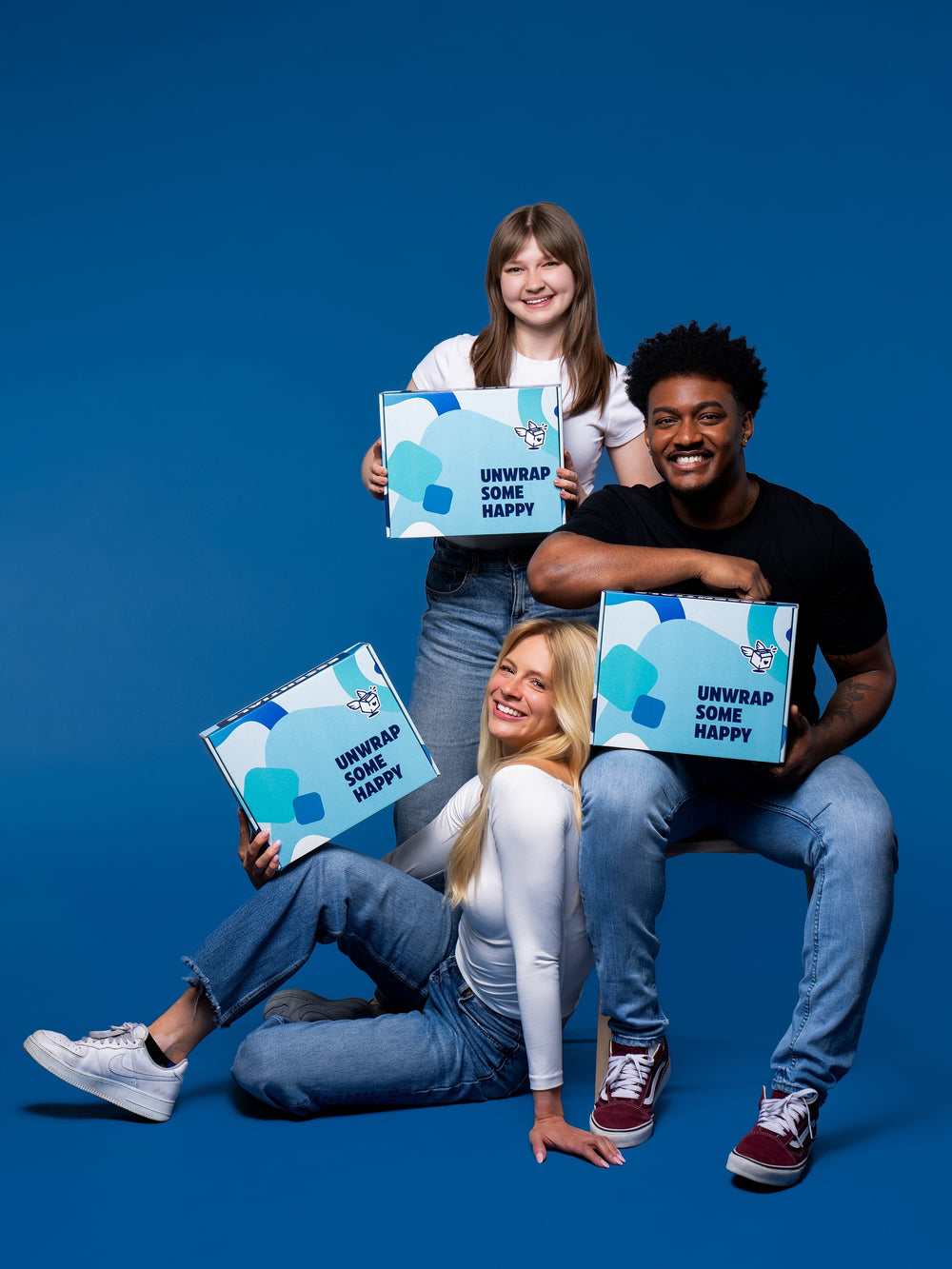 Three smiling young people hold blue and white boxes labeled "unwrap some happy" against a bright blue background.
