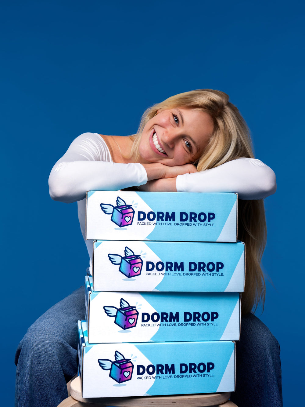 Smiling blonde woman hugging a stack of four blue and white Dorm Drop boxes against a blue background.