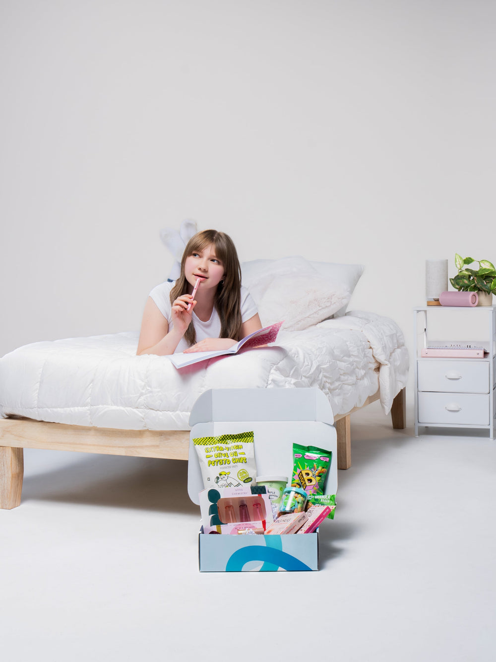 A girl on a bed reads while a Dorm Drop care package filled with snacks sits on the floor in front of her.