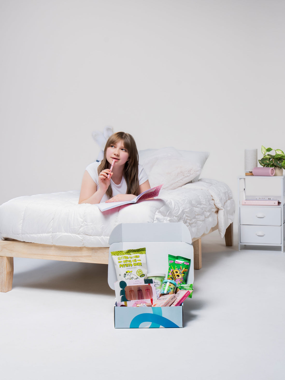 A girl on a bed reads while a Dorm Drop care package filled with snacks sits on the floor in front of her.