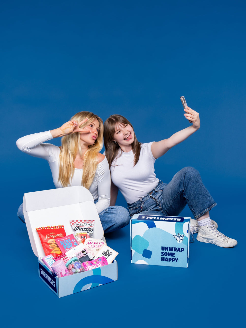 Two young women take a selfie against a blue background next to open care packages filled with snacks and products.