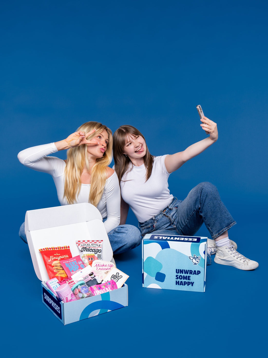 Two young women take a selfie against a blue background next to open care packages filled with snacks and products.