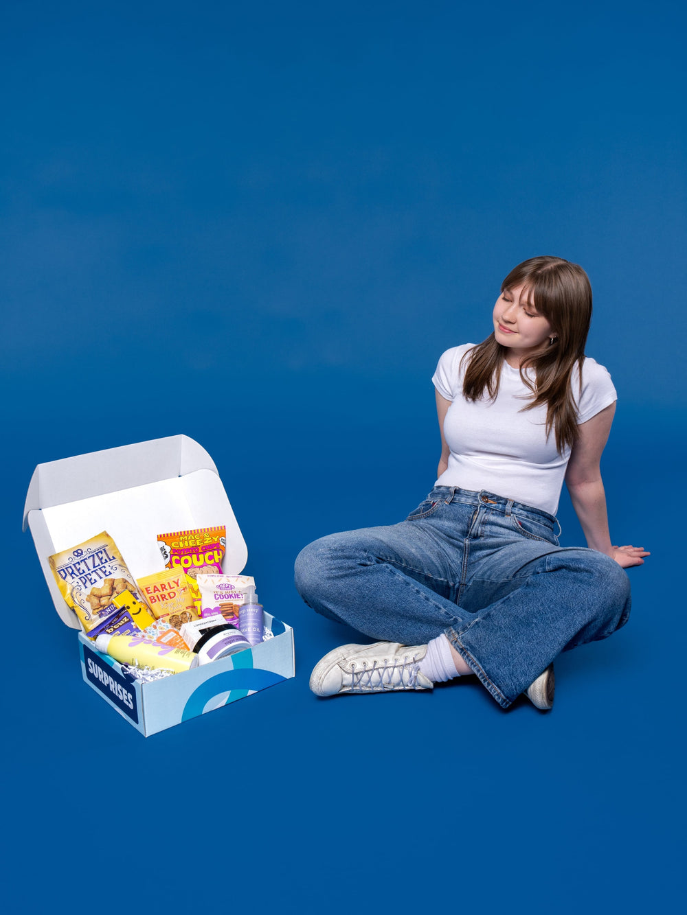 A young woman sits on a blue background next to an open white care package box filled with snacks and items.