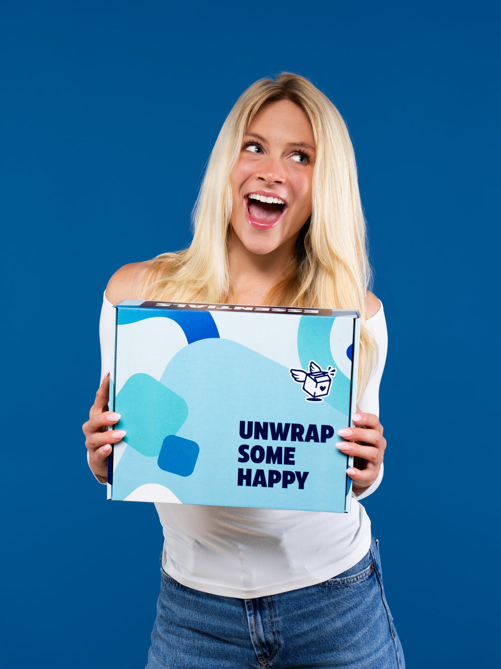 A smiling woman holds a light blue Dorm Drop care package box printed with abstract shapes and "unwrap some happy" text.