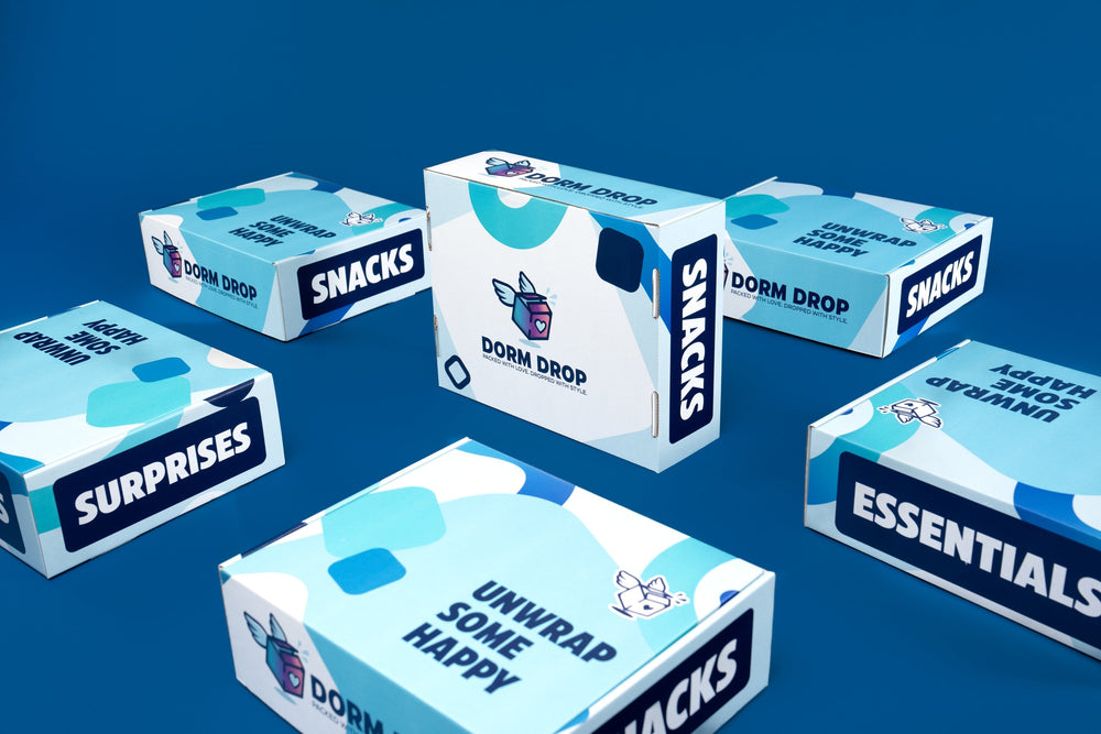 Five white and turquoise Dorm Drop boxes labeled Snacks, Surprises, and Essentials arranged on a blue background.