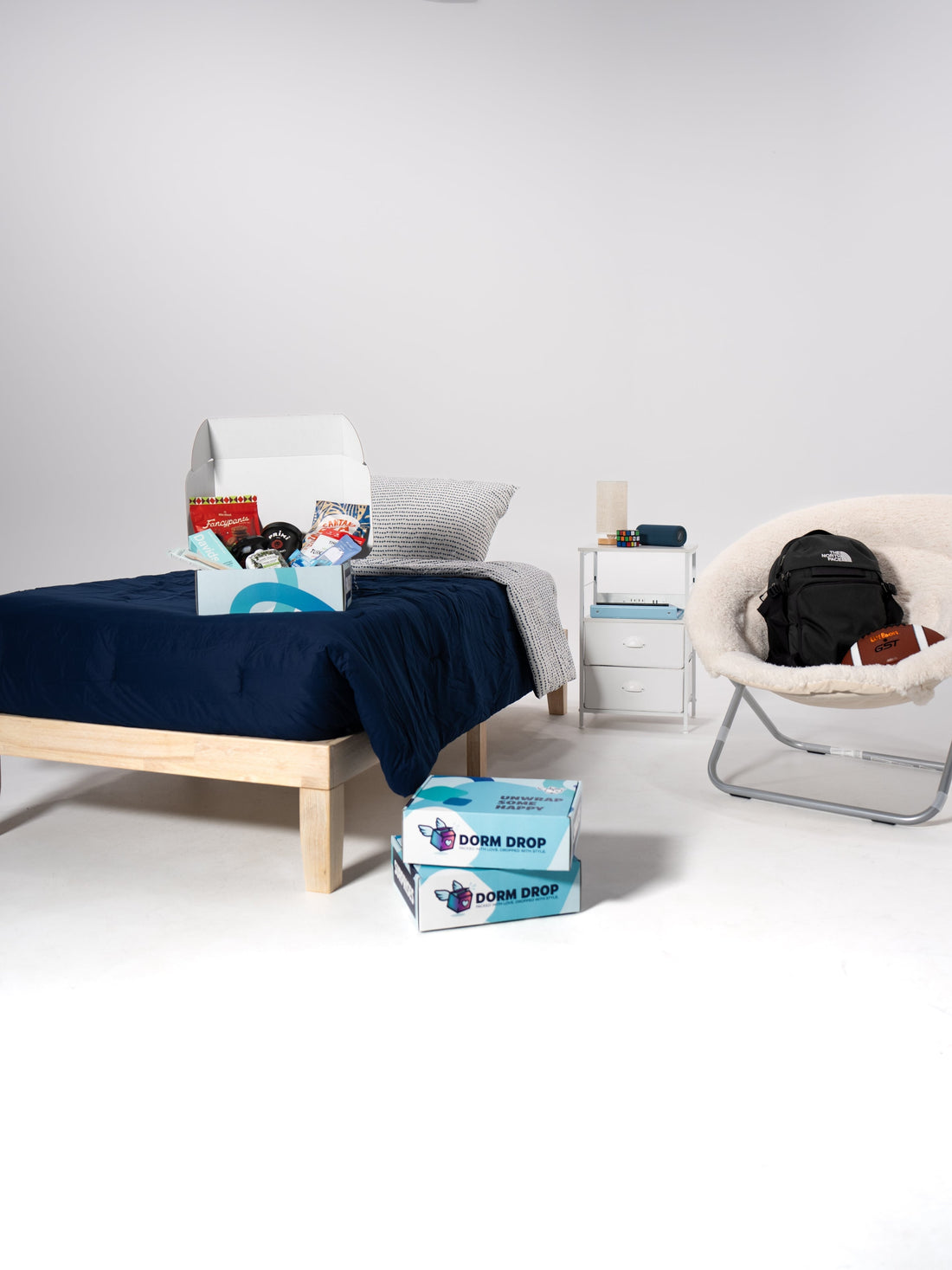 A dorm room display with a wooden bed, navy bedding, nightstand, papasan chair, and two stacked Dorm Drop boxes on the floor.