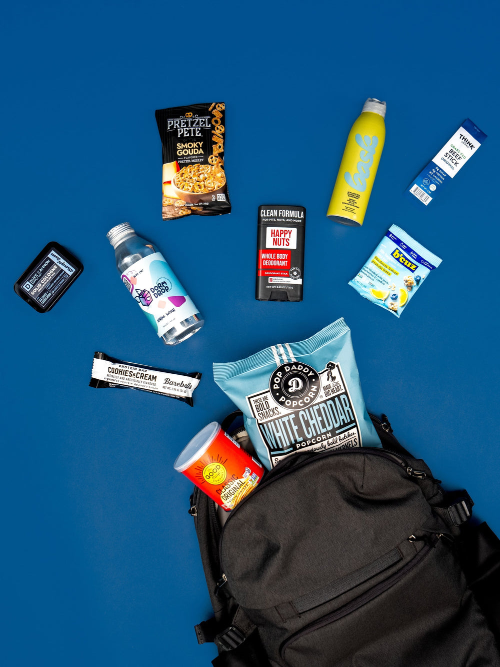 A black backpack overflowing with snacks and personal care items on a bright blue background.