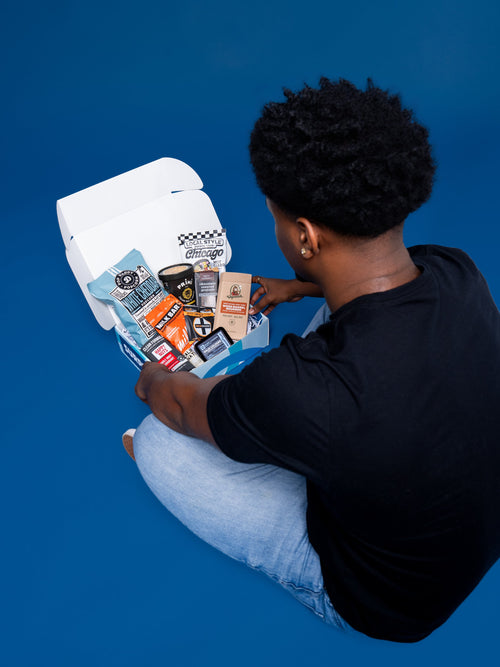 A college student in a black tee opens a Dorm Drop care package box, overhead view