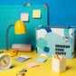 A closed Dorm Drop Essentials box reading “Unwrap Some Happy” on a student desk with a lamp, alarm clock and sticky notes