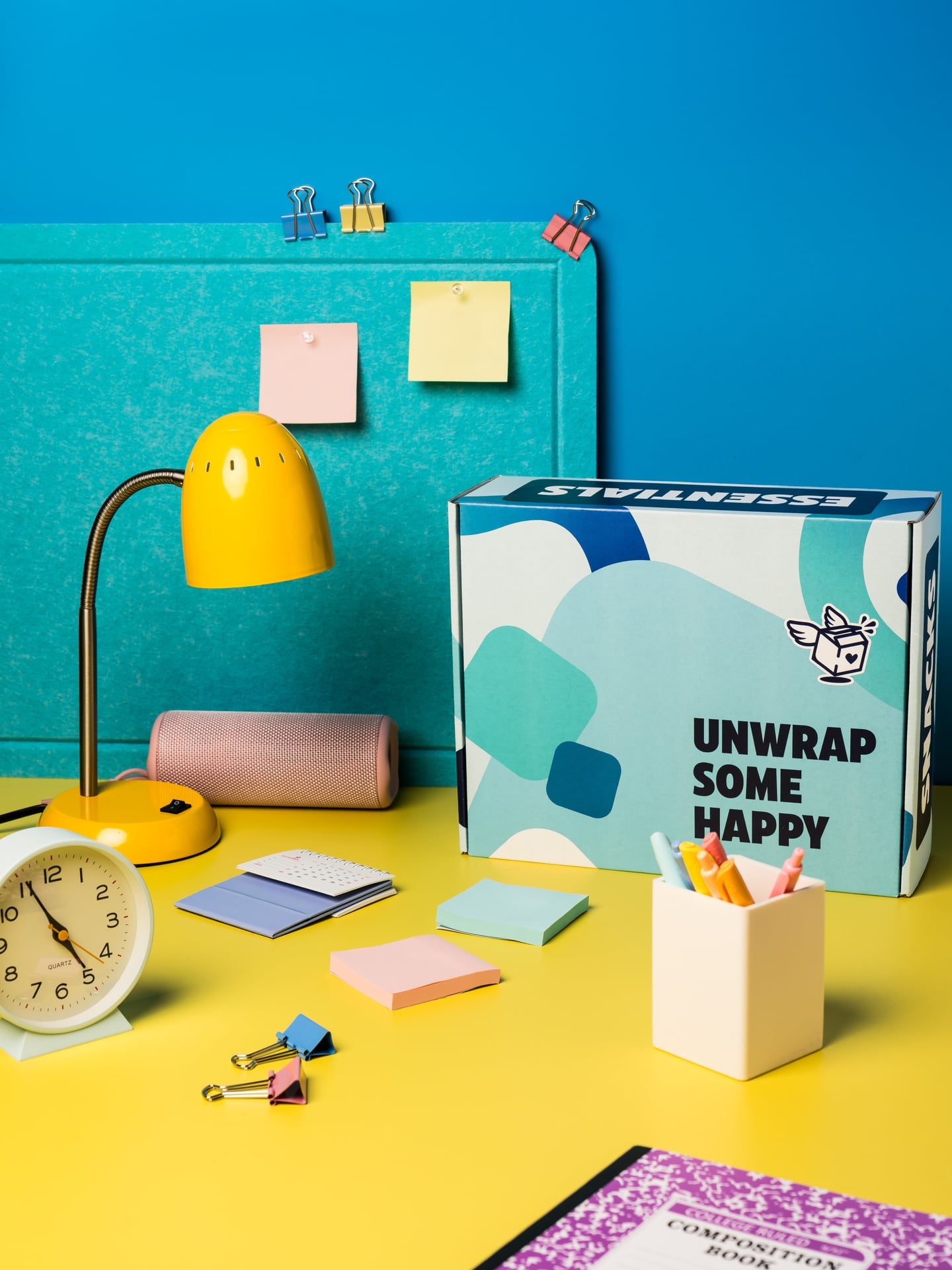 A closed Dorm Drop Essentials box reading “Unwrap Some Happy” on a student desk with a lamp, alarm clock and sticky notes